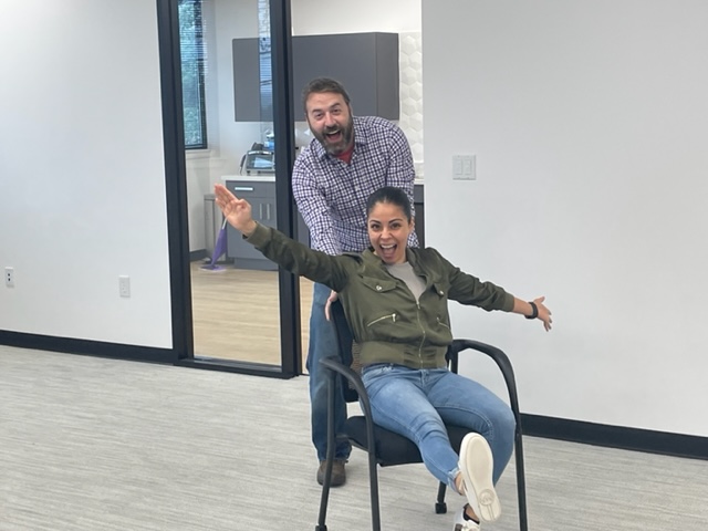Two people in an office: a woman sits in a chair with her arms outstretched in a playful pose, while a man playfully pushes the back of the chair.